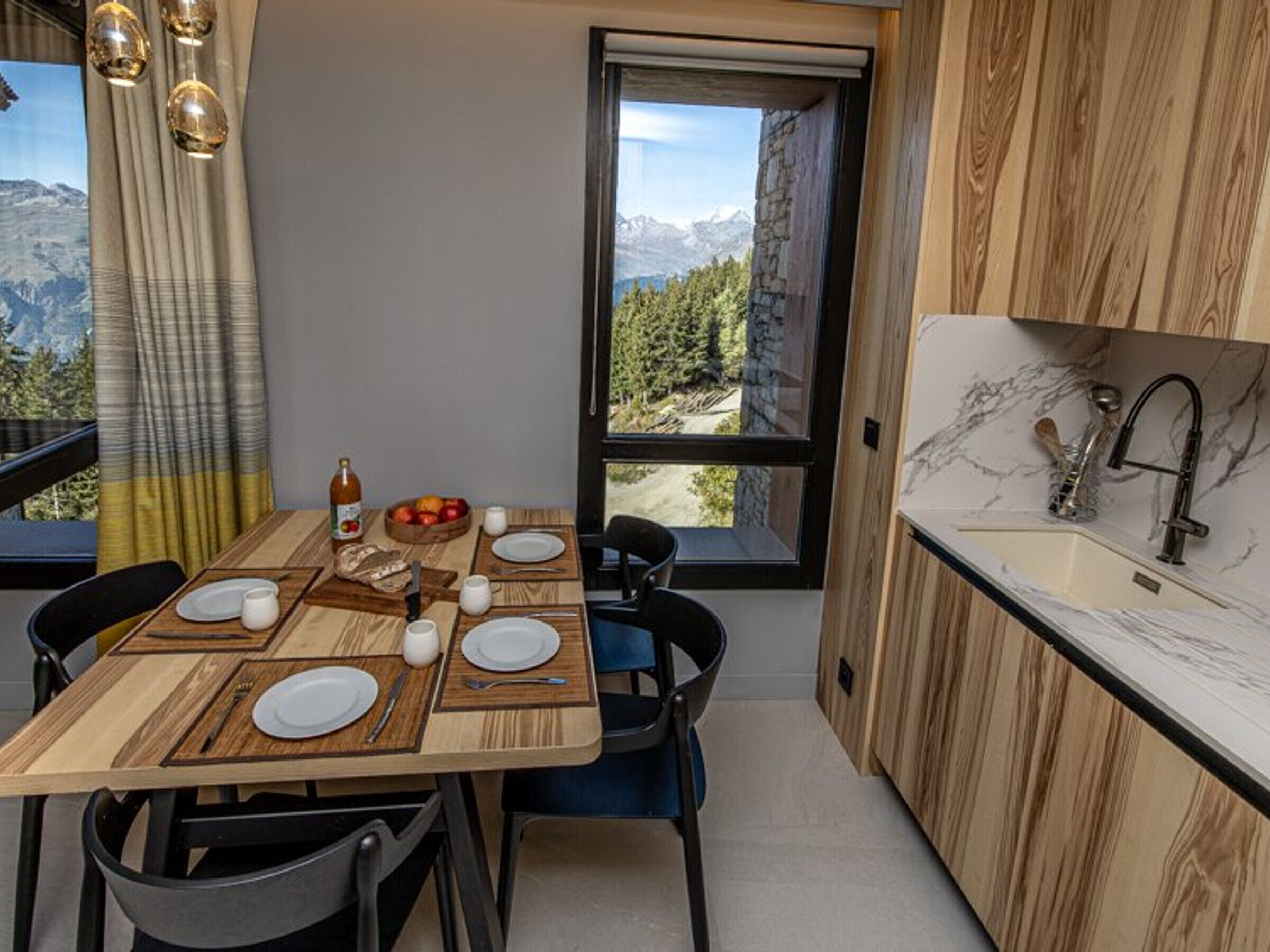 Eine moderne Küche mit einem Holztisch, gedeckt für vier Personen, einer Spüle mit Marmorarbeitsplatte und Blick auf Berge durch ein großes Fenster.