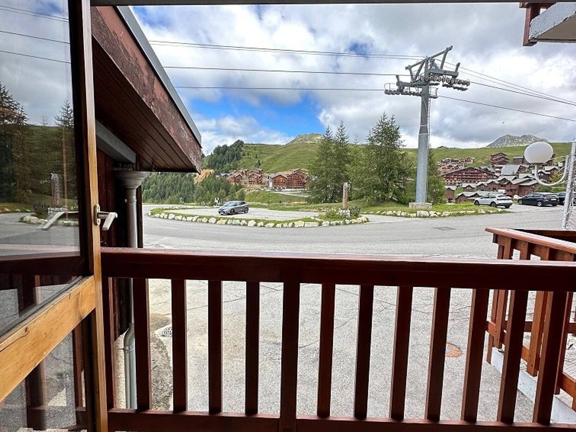 Vista da una finestra con ringhiera in legno, su un'area di parcheggio con alcune auto, un edificio con tetto spiovente e un paesaggio montuoso sullo sfondo.
