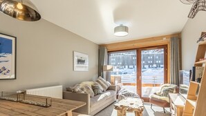 A cozy living room with a wooden table, a sofa, and a view of a snowy landscape through large windows.