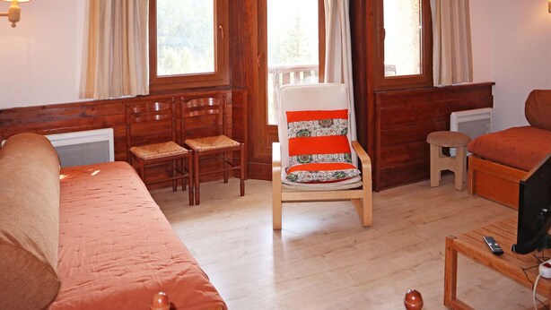 Una habitación acogedora con suelo de madera, un sofá, un sillón con un cojín estampado, una mesita con televisor y una ventana con cortinas.