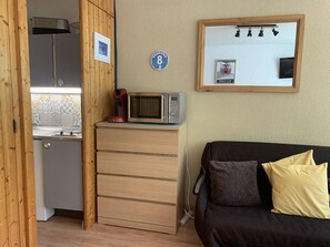 Eine kleine Küchenzeile mit Mikrowelle, Kaffeemaschine und kleinem Kühlschrank. Es ist ein Holzschrank mit Schubladen sowie eine schwarze Couch mit gelben Kissen zu sehen. Darüber befindet sich ein Spiegel mit Beleuchtung und ein Schild mit der Nummer 8.