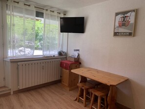 A small kitchen with a wooden table and stools, a radiator, a window with curtains, a television on the wall, and a poster on the wall. - Cozy studio near slopes at Plagne Centre, 4 pers. (La Plagne-Tarentaise)