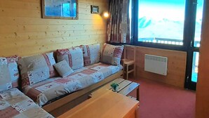 Une pièce chaleureuse avec un mur en bois, un canapé agrémenté de coussins à motifs, une petite table et une fenêtre donnant sur une montagne.