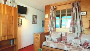 Un salon chaleureux avec un canapé en bois, un téléviseur fixé au mur et une fenêtre ornée de rideaux à motifs.