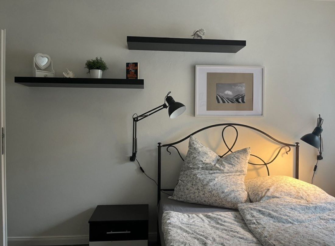 A bedroom with a bed, bedside lamps, a nightstand, a framed picture, and two wall-mounted shelves with decorative items.
