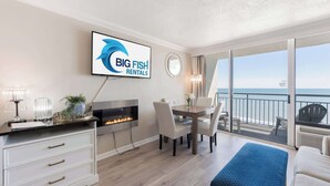 Une chambre moderne avec une cheminée, une table à manger avec des chaises, un téléviseur diffusant le programme « BIG FISH RENTALS », et un balcon offrant une vue sur l’océan.
