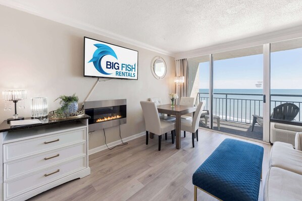 A modern room with a fireplace, a dining table with chairs, a TV displaying 'BIG FISH RENTALS,' and a balcony with a view of the ocean.