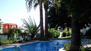 A swimming pool surrounded by palm trees and greenery, with a person sitting on a bench and another person swimming.