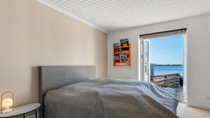 A bedroom with a large bed, a bedside table with a lamp, a bookshelf, and a view of the sea through an open door.