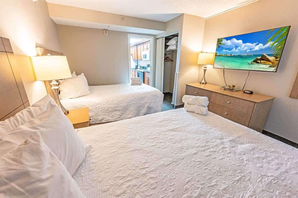 A hotel room with two beds, a TV, a desk, and a view of the outside.