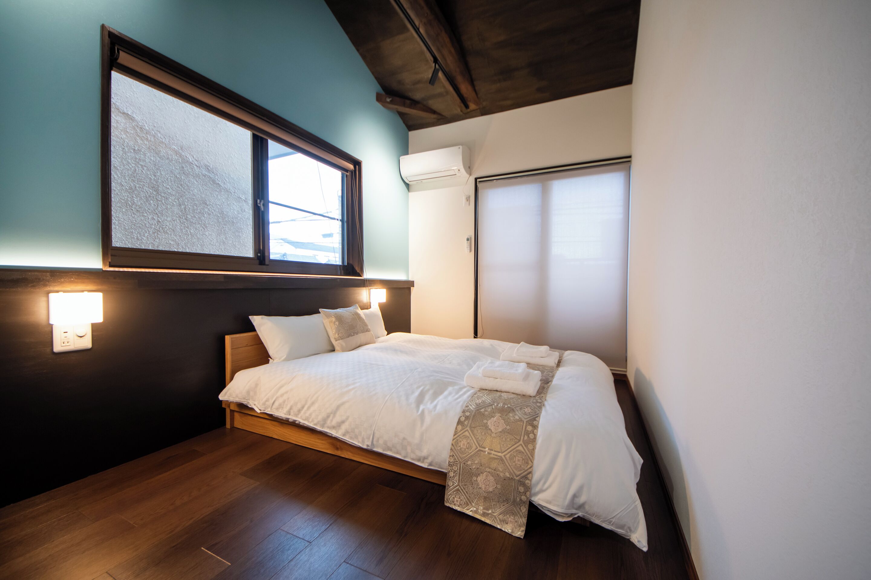 A bedroom with a bed, a window, an air conditioning unit, and a wall-mounted light fixture.