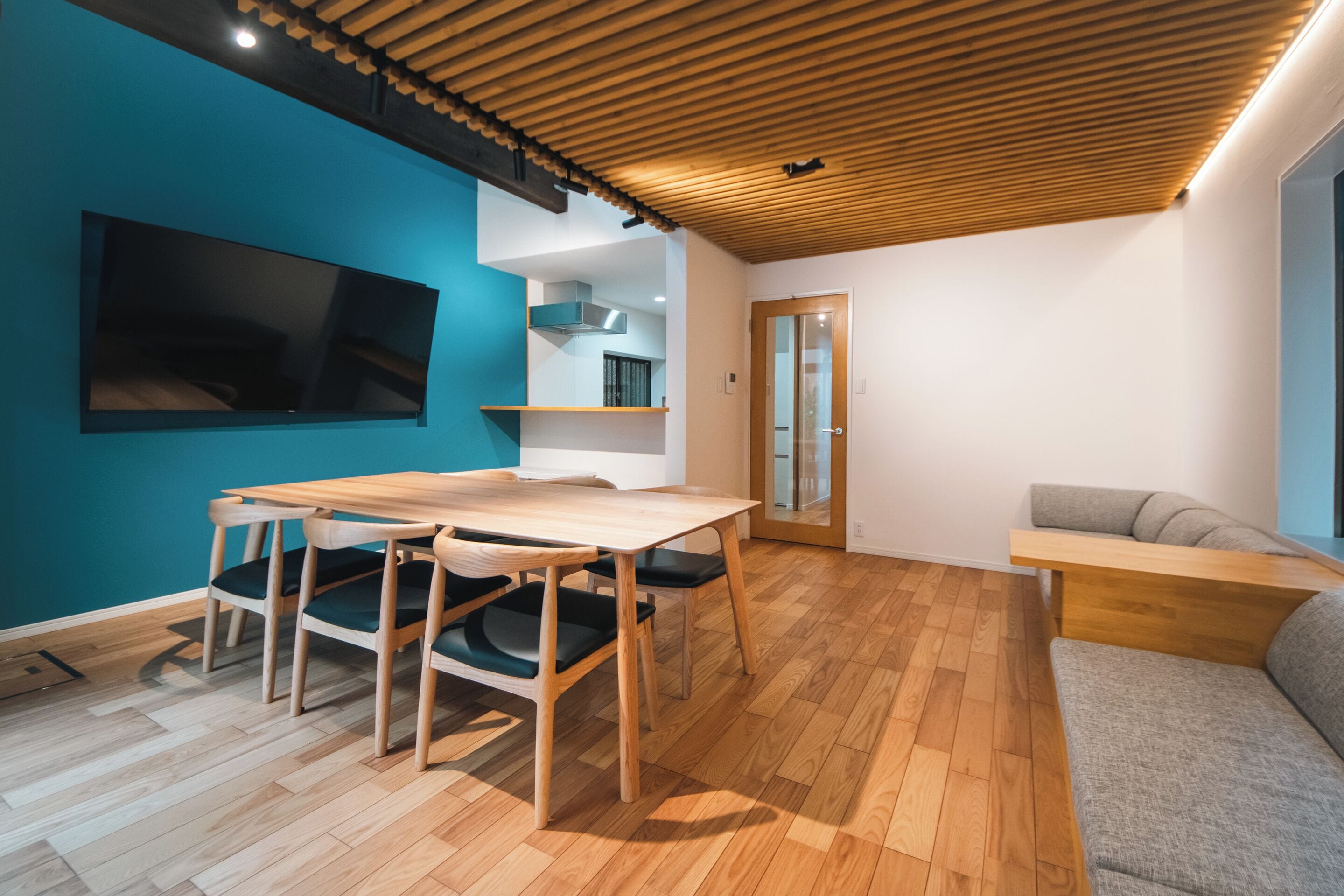 Un interior modern amb un sostre de fusta, una paret d'accent blau, una zona de menjador amb taula i cadires, i un sofà gris.