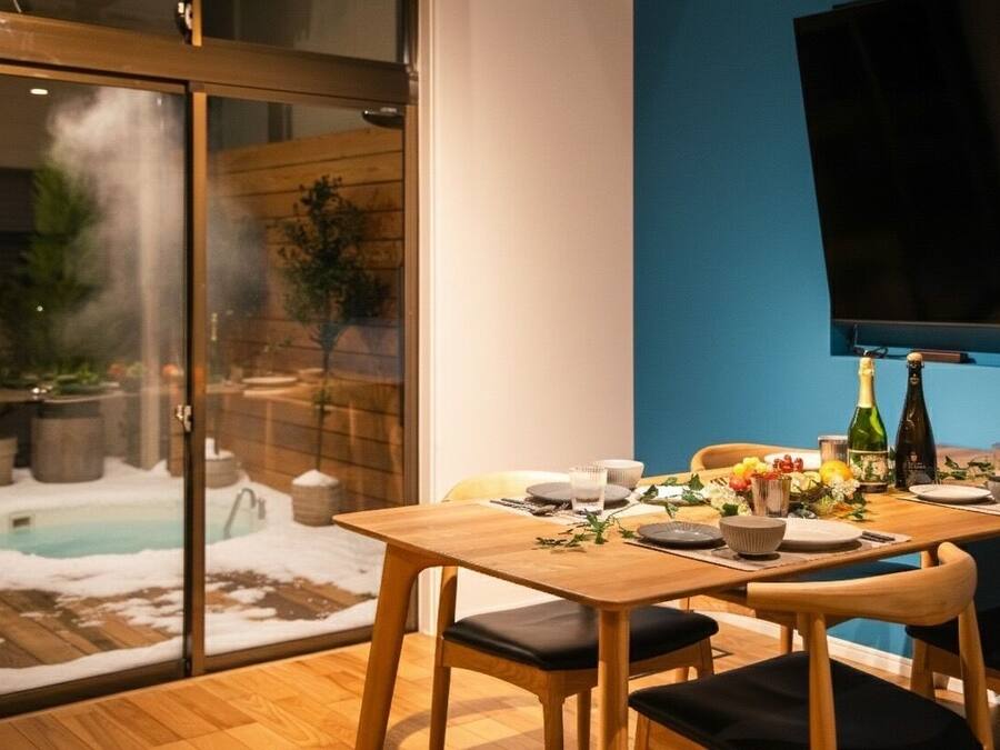 A dining area with a table set for two, a TV mounted on the wall, and a view of a pool through a glass door.