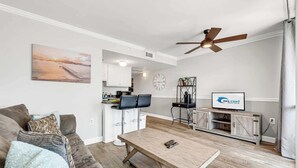 A living room with a sofa, coffee table, and a TV. There's a kitchen area with cabinets and a clock. A ceiling fan is visible. - Meridian Plaza 308 - 1 Bedroom (Myrtle Beach)