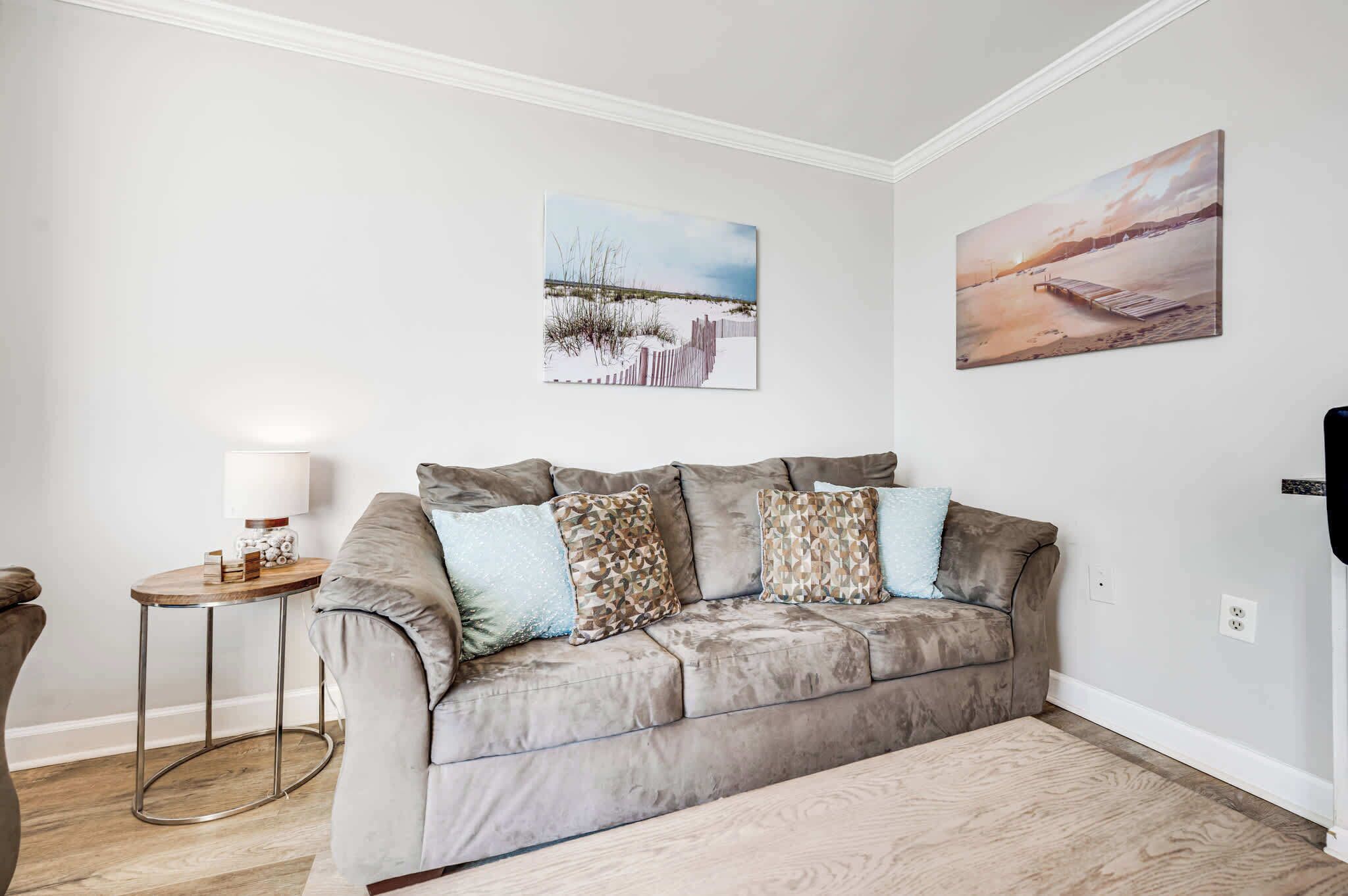 Een woonkamer met een beige bank, twee aan de muur hangende schilderijen, een klein houten bijzettafeltje met een lamp, en een lichtkleurig vloerkleed.