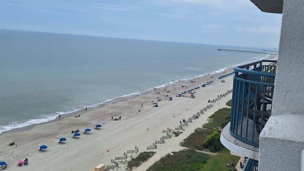 Ein Strand mit Sonnenschirmen und Personen, ein Steg in der Ferne und ein Gebäude mit einem Balkon.
