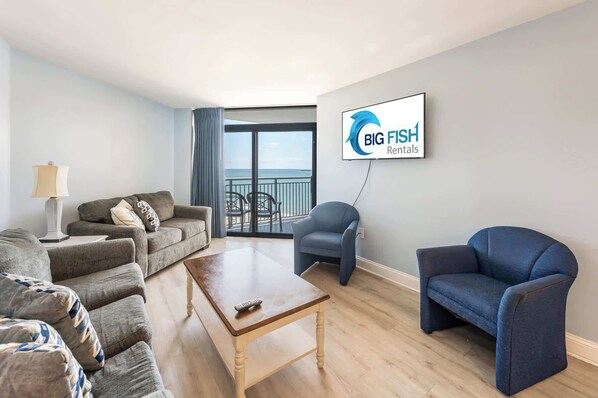 A living room with a sofa, armchair, and coffee table, a TV mounted on the wall, and a balcony with a view. - 3BR in Sand Dunes Resort, Oceanview, King Suite! (Myrtle Beach)