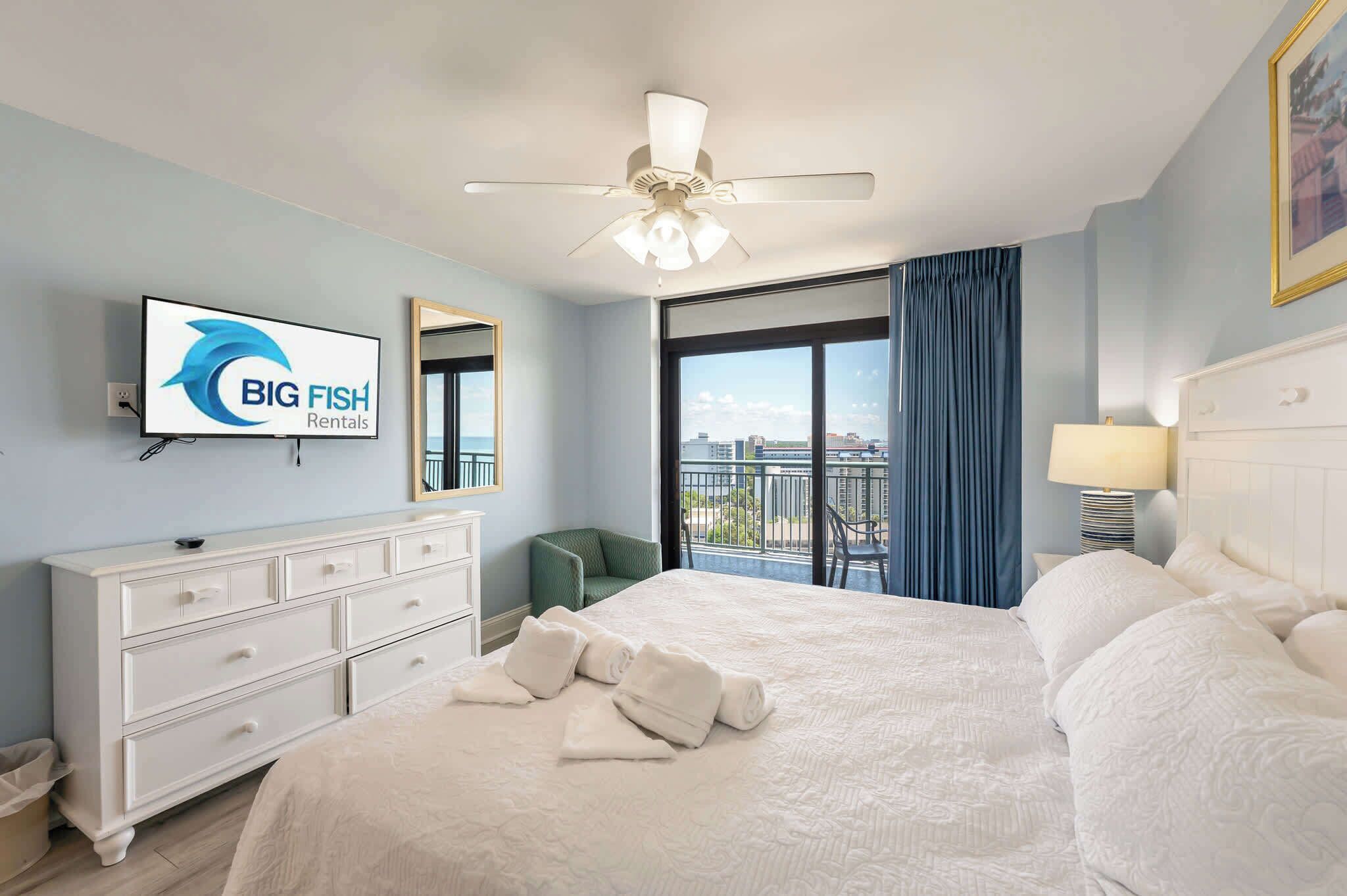 A hotel room with a bed, a ceiling fan, a TV displaying 'BIG FISH Rentals,' a balcony with a view, and a green chair.