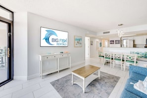 Uma sala de estar moderna com uma mesa de consola branca, uma mesa de centro de madeira, um sofá azul e uma área de cozinha com armários brancos e uma mesa de jantar.