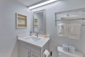 Ein Badezimmer mit einem weißen Waschbecken, Spiegel, Handtuchhalter, Toilettenpapier und Ablage mit Pflegeprodukten.