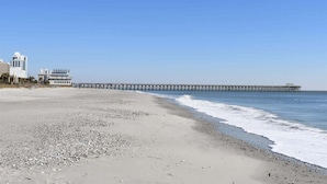 A beach with a long pier extending into the ocean, a building with a white facade, and a clear blue sky.