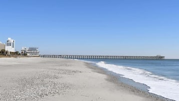 A beach with a long pier extending into the ocean, a building with a white facade, and a clear blue sky.