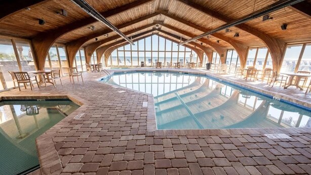 An indoor swimming pool with a brick floor and wooden ceiling, surrounded by tables and chairs.