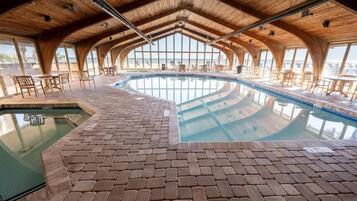 An indoor swimming pool with a brick floor and wooden ceiling, surrounded by tables and chairs.