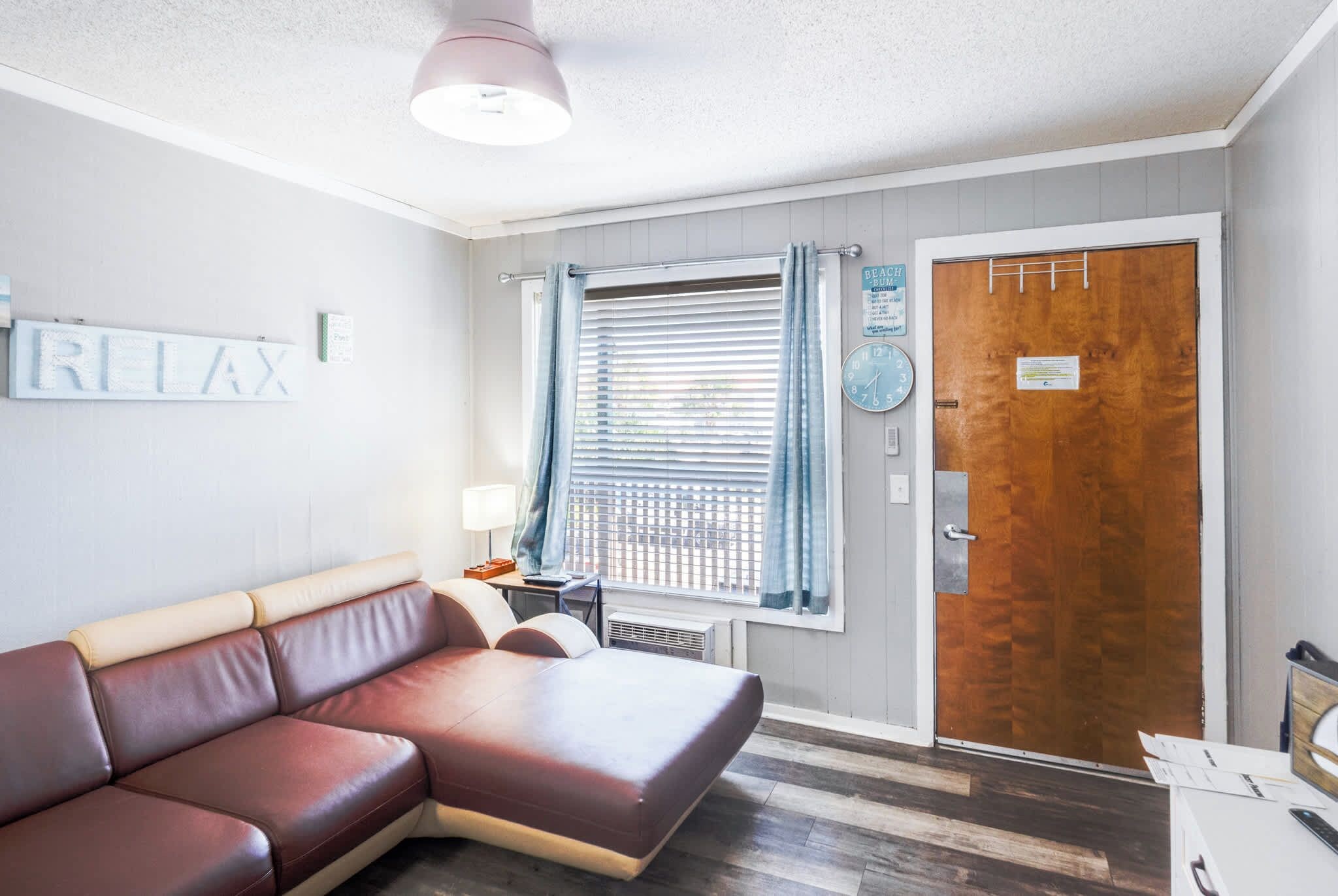 A room with a brown leather sofa, a wooden door, a window with blinds, and a wall-mounted clock.