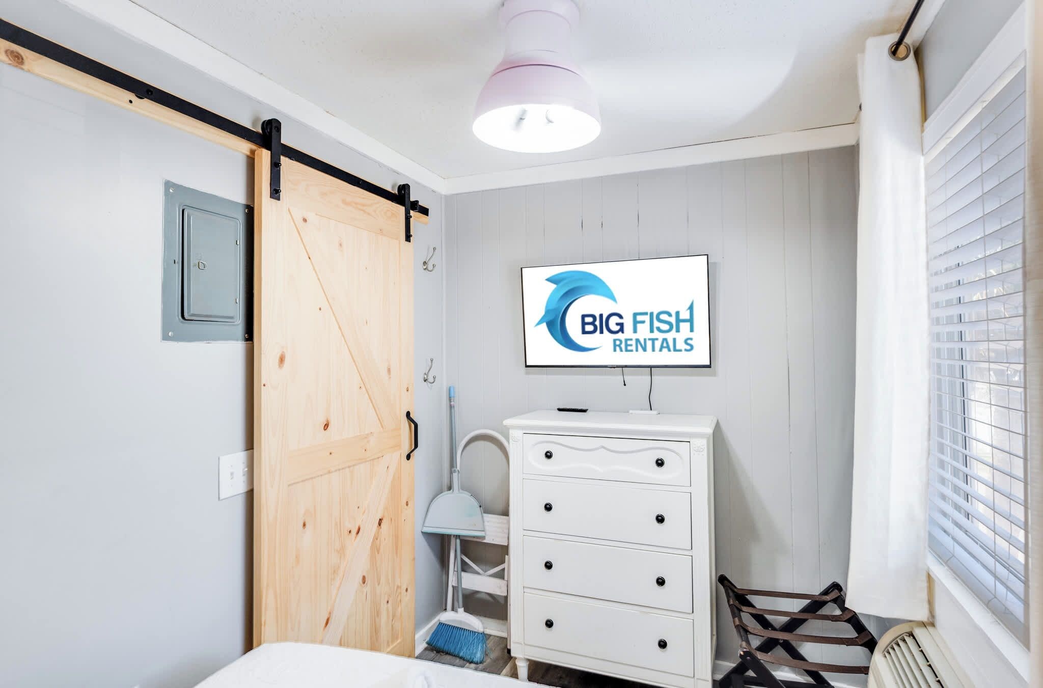 Un dormitorio con puerta corredera, cómoda, televisor mostrando 'BIG FISH RENTALS' y ventana con persianas.