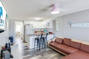A modern kitchen with white cabinets, a refrigerator, and a pink pendant light. There is a dining area with a brown leather couch and two black chairs. - 1BR Sea Villas in Windy Hill - Steps to the Beach! (North Myrtle Beach)