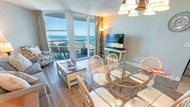 A modern living room with a glass dining table, comfortable seating, a flat-screen TV, and a view of the ocean. - Coastal condo with hot tub & pools (North Myrtle Beach)