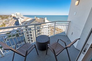 Een balkon met twee rieten stoelen en een ronde tafel, met uitzicht op een strand en de oceaan.