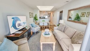 Una sala de estar moderna con un sofá, una mesa de centro y un televisor. Se observa un comedor con una mesa y sillas. La cocina se ve al fondo.