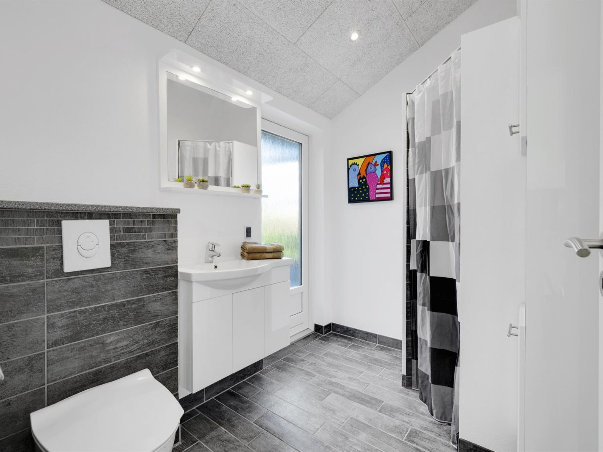 Ein modernes Badezimmer mit Waschbecken, Spiegel und Toilette in Weiß. Eine Duschvorhang und ein kleines Fenster mit Blick auf Grünfläche.