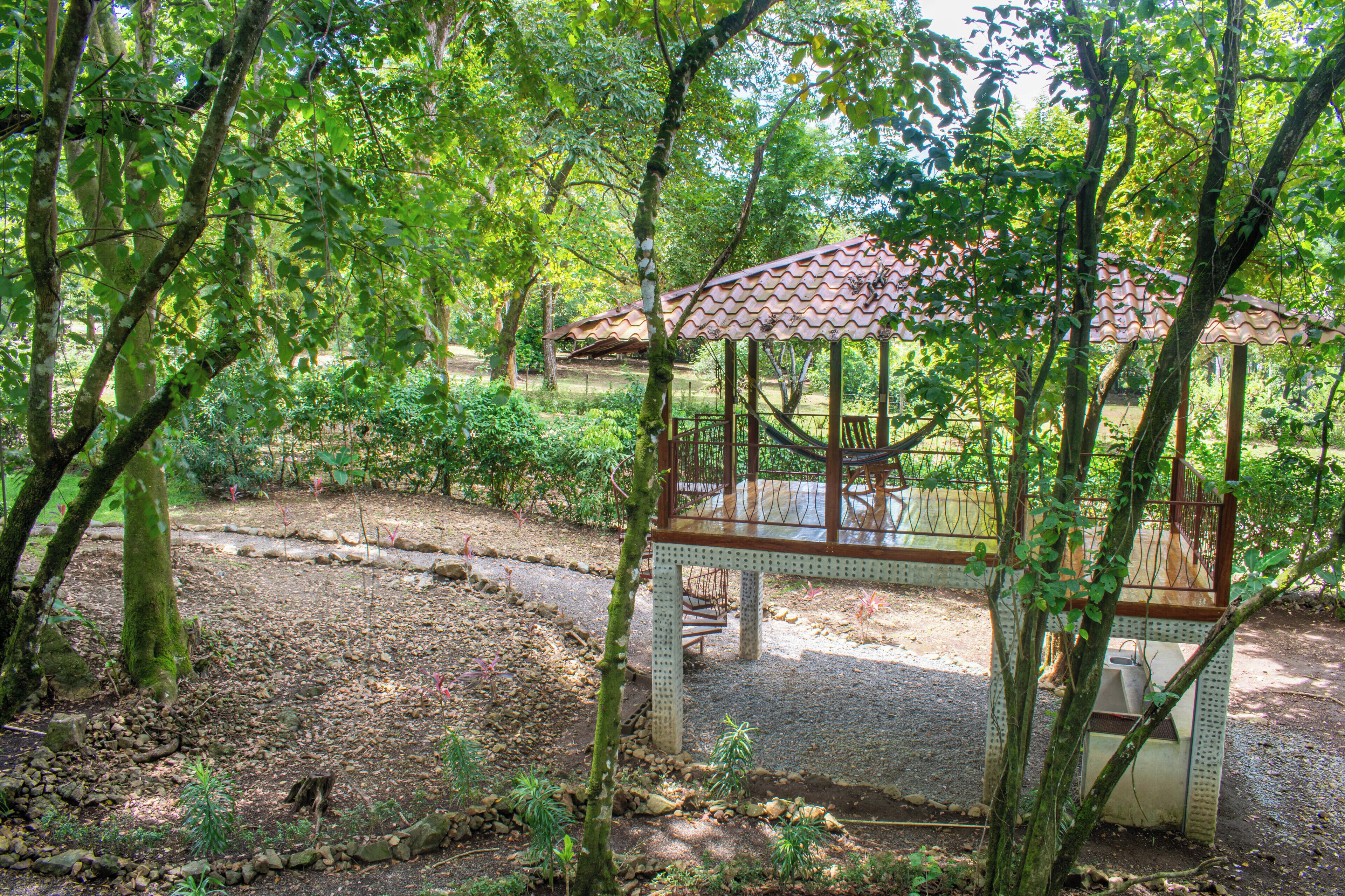 A gazebo with a tiled roof, surrounded by trees and a gravel path.