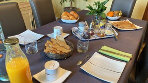 Una mesa de comedor con cruasanes, pan, un frutero, un jarrón con flores y una botella de jugo de naranja.
