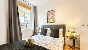 Habitación de hotel con cama, ventana con cortinas, mesita de noche con lámpara y un cuadro en la pared.