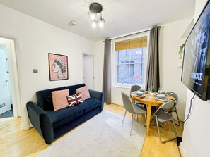 A living room with a dark blue sofa, a dining table with chairs, a television, and a window with curtains. - Cozy 1-Bedroom flat with Wi-Fi, Washer/Dryer near Shoreditch, Old Street, Hoxton (London)