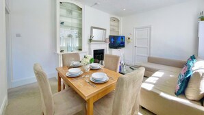 Un comedor con una mesa de madera preparada para cuatro, un aparador de vidrio con vajilla, un televisor en la pared y un sofá con cojines decorativos.