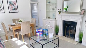 Una sala de estar con una mesa de centro de vidrio, sillas color beige, una mesa de comedor con platos y vasos, una chimenea y una estantería.
