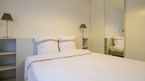 A neatly made bed with white bedding and a beige accent, a bedside table with a lamp, and a mirror on the wall.