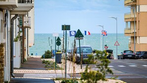 A coastal street with parked cars, a pedestrian crossing, and flags of France, the UK, and another country.