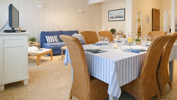 A dining area with a table set for four, surrounded by wicker chairs, a sofa, and a television in the background.