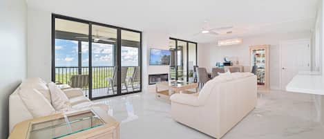 A spacious living room with a large sliding glass door leading to a balcony, a flat-screen TV mounted on the wall, a white sofa, and a glass coffee table.