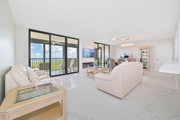 A spacious living room with a large sliding glass door leading to a balcony, a flat-screen TV mounted on the wall, a white sofa, and a glass coffee table.