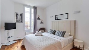Een slaapkamer met een bed, een televisie, een raam met gordijnen, een lijst met een afbeelding en een spiegel aan de muur.