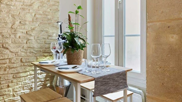 Eettafel gedekt met borden, glazen en een potplant, geplaatst bij een raam met een stenen muur op de achtergrond.