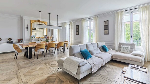 A spacious living room with a large sectional sofa, a dining area with a wooden herringbone floor, and large windows offering a view of greenery. - Incredible Apartment  Etoile Porte Maillot (Paris)