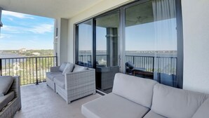 Een balkon met rieten meubels en een witte bank, met uitzicht op de zee en gebouwen.